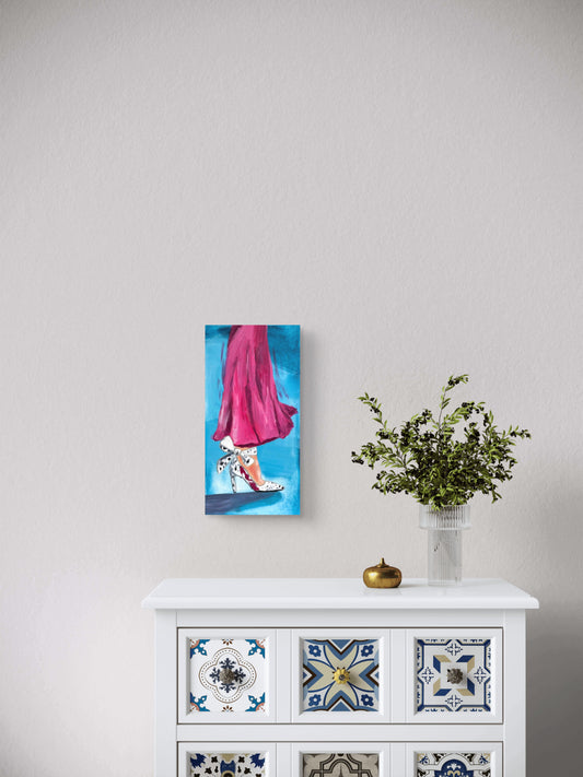 Painting of fashion shoe and a pink dress hung over a vase of greenery on a light gray wall.
